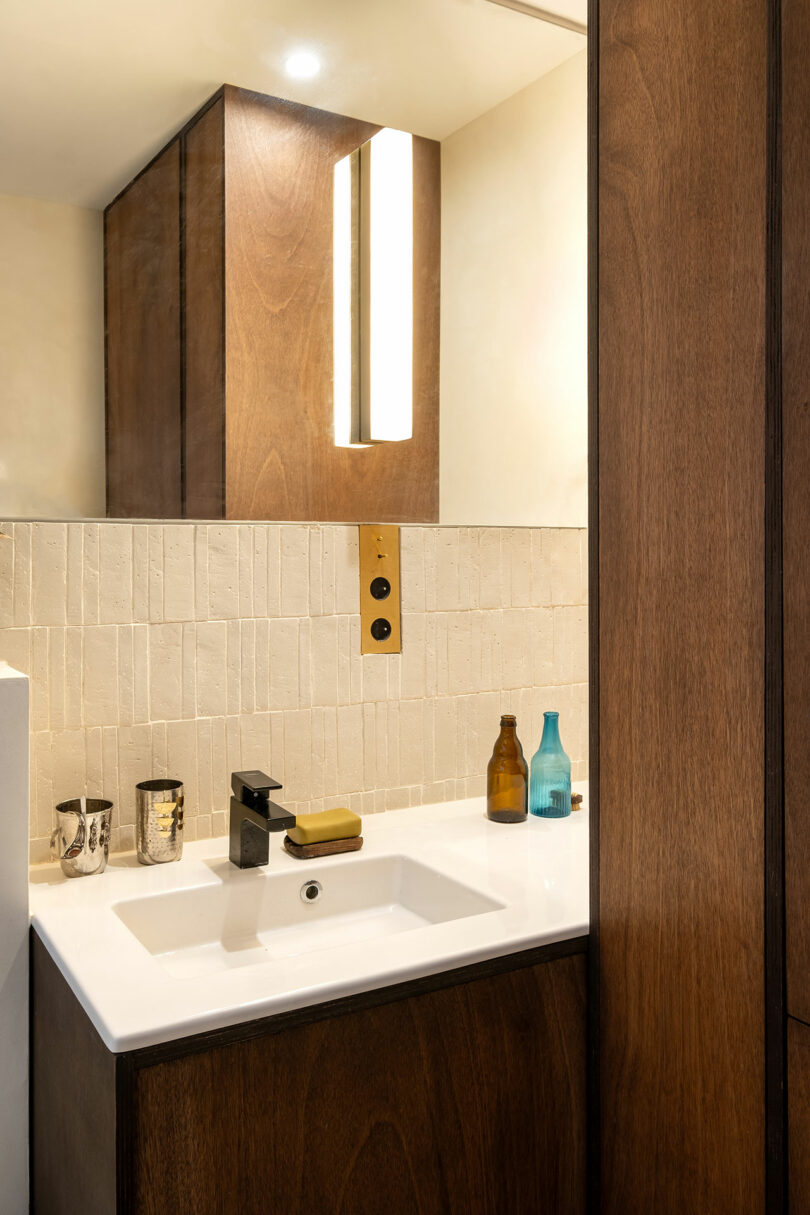 Modern bathroom sink with a rectangular white basin, black faucet, soap, and bottles on the counter. Vertical wall light and wooden cabinetry are visible in the background.