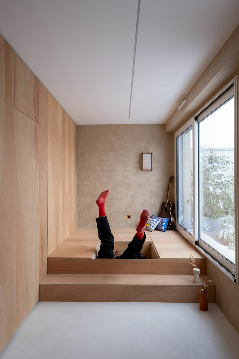 Person with red socks lies on their back with legs up in a minimalist wooden room, next to a window, a guitar, and a low table, with natural light coming in.