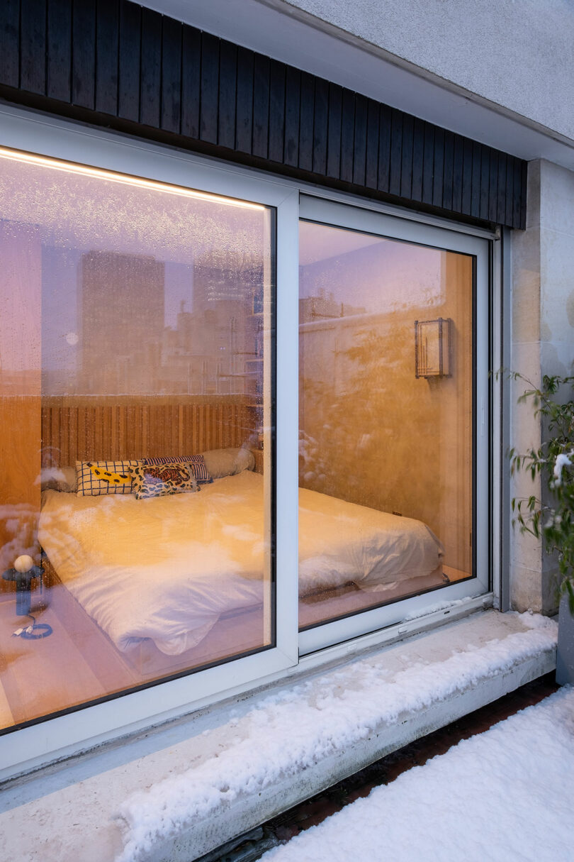 A cozy bedroom with a double bed and patterned pillows is seen through large glass doors, with snow covering the ground and window ledge outside.