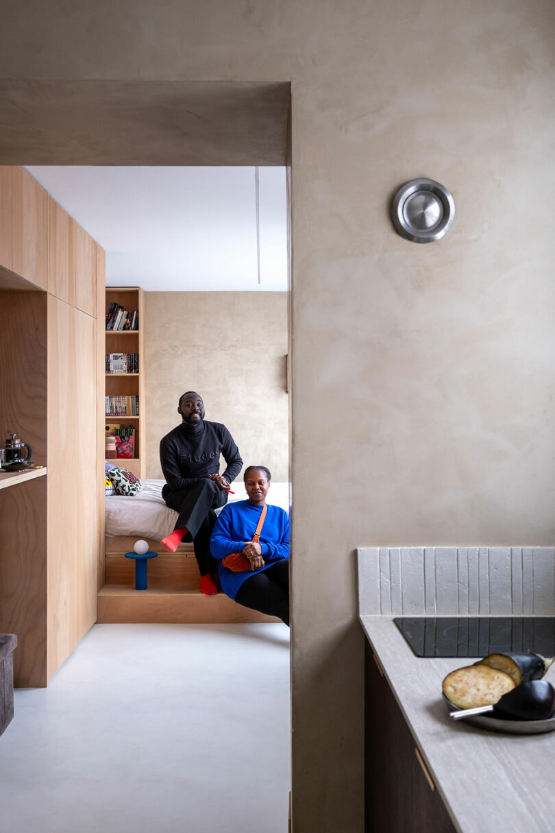 Two people sit together in a minimalist, beige-toned apartment; one on a raised bed, the other on the step below. Kitchen counter with eggplants is visible in the foreground.