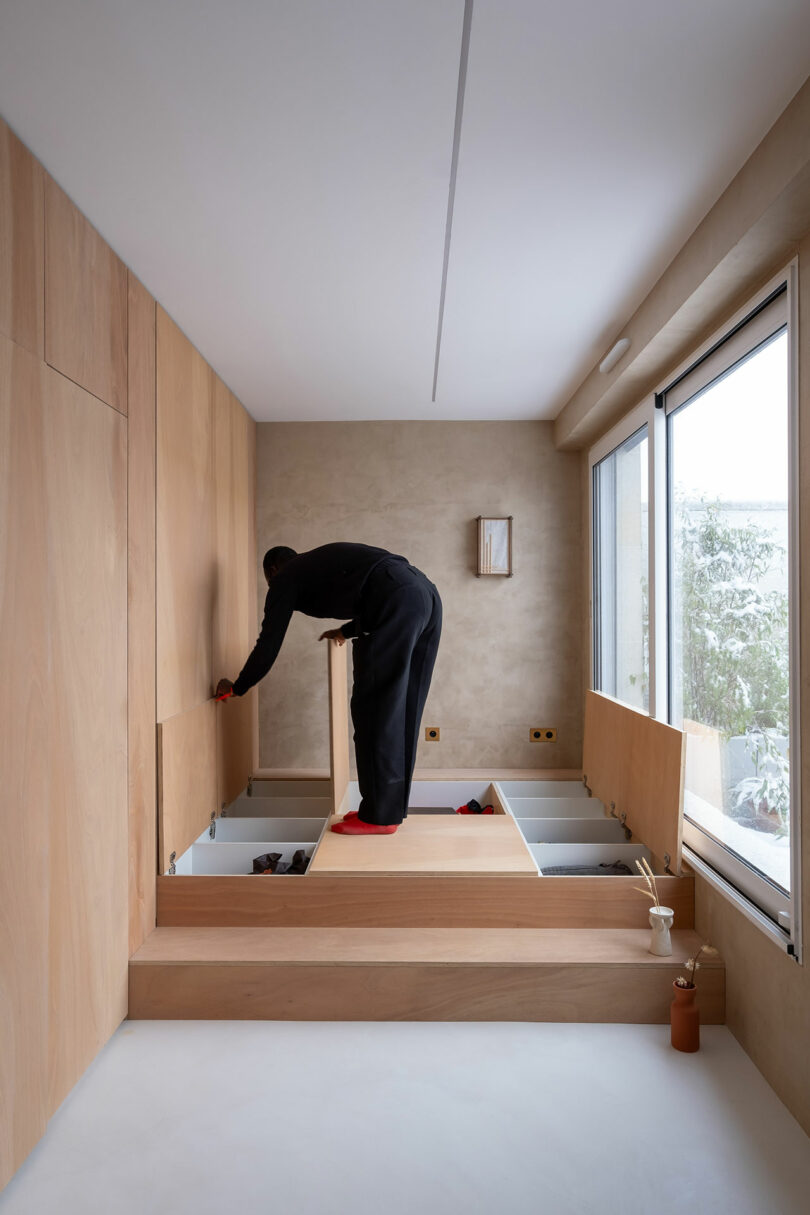 A person stands on a wooden platform with built-in storage compartments, opening one lid in a minimalist room with large windows and light wood paneling.