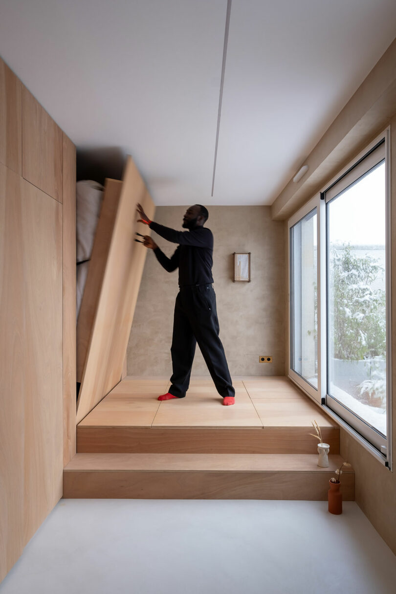 A person lifts a foldaway bed integrated into a wooden platform in a minimalist, modern room with natural light.