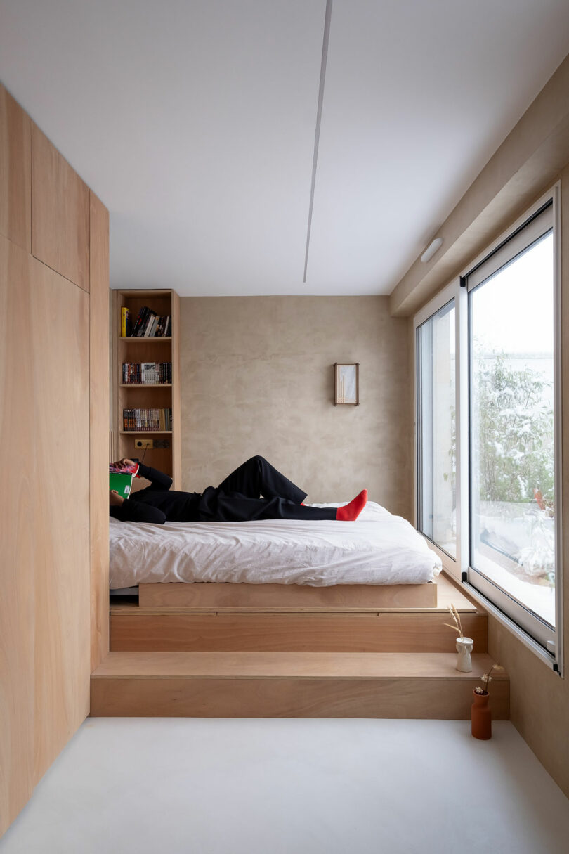 Person lying on a raised bed reading a book in a minimalist bedroom with light wood paneling, built-in shelves, large window, and neutral-colored walls.