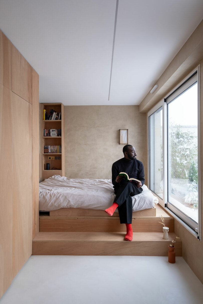 A man with red socks sits on the edge of a bed in a minimalist bedroom with wooden accents, holding a book and looking out a large window.
