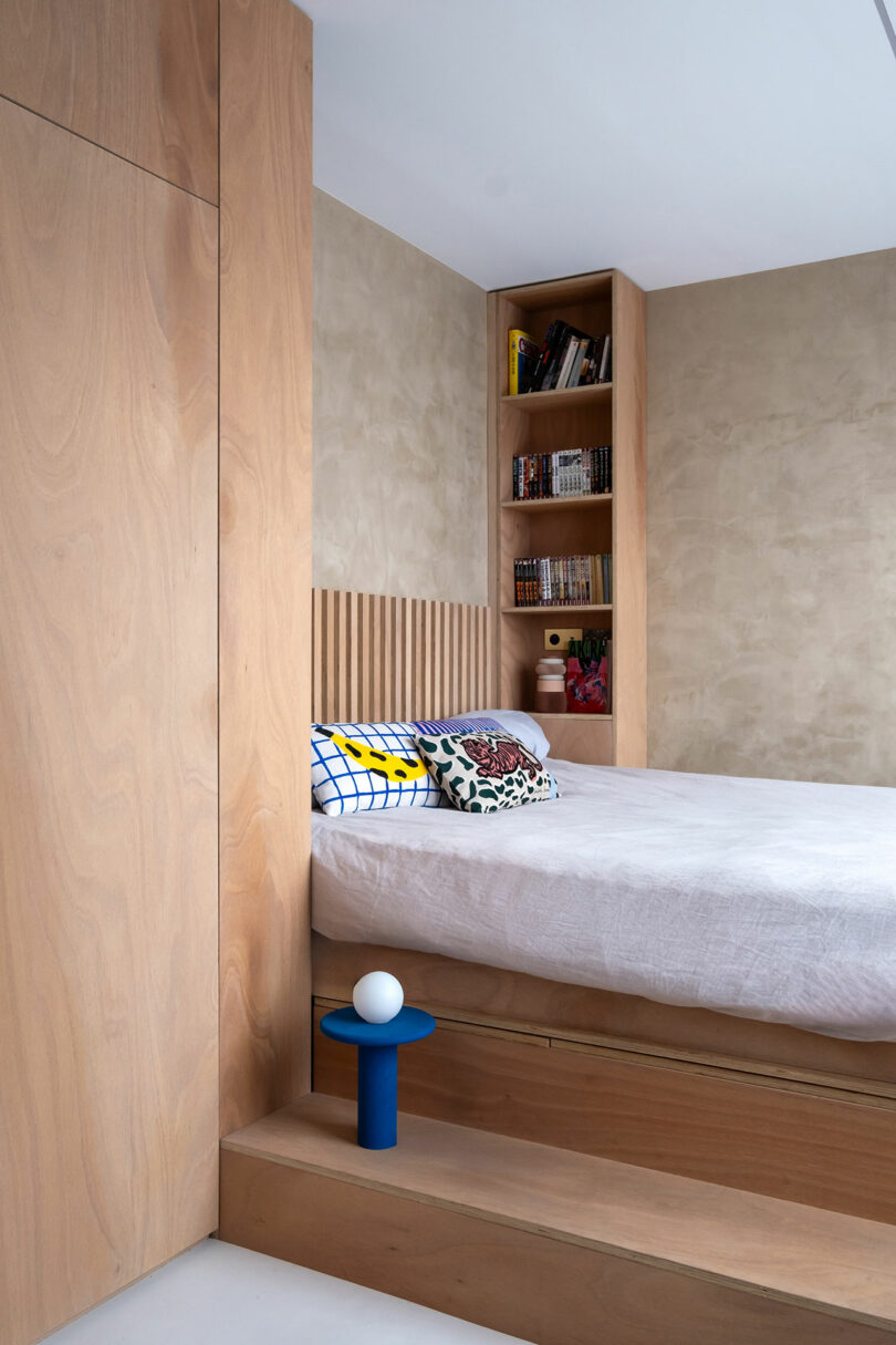 A modern bedroom with light wood paneling, a built-in bookshelf, a bed with patterned cushions, and a small blue nightstand with a spherical lamp.