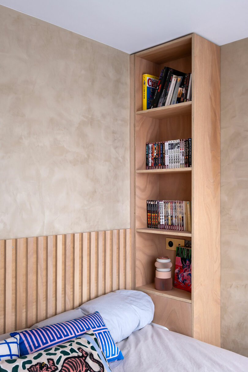 Wooden bookshelf built into a bedroom wall above the bed, holding books, manga, and small decorative items near a slatted wooden headboard.