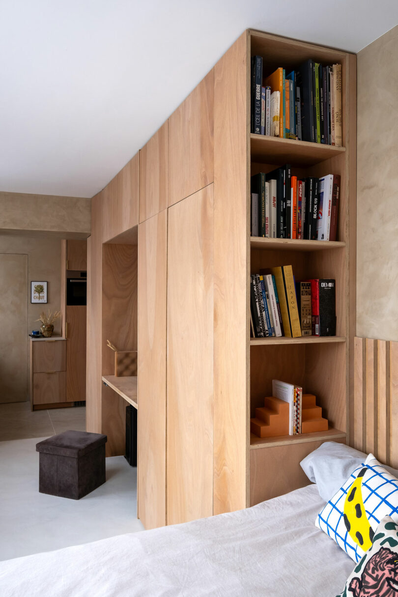 A modern bedroom features light wood built-in shelves and cabinetry, with books and decor items, adjacent to a bed with patterned pillows.