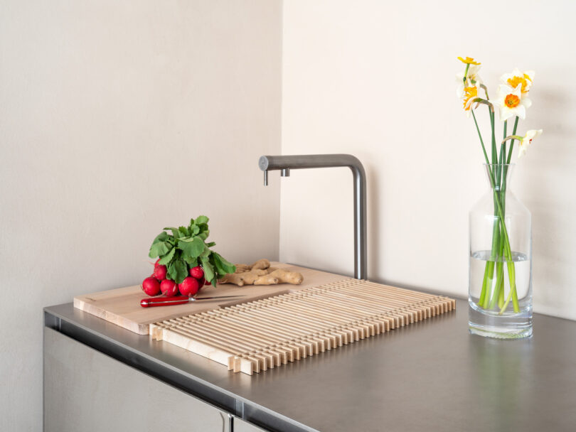 A modern kitchen countertop with a vase of daffodils, a faucet, ginger roots, radishes, and a knife on a wooden board