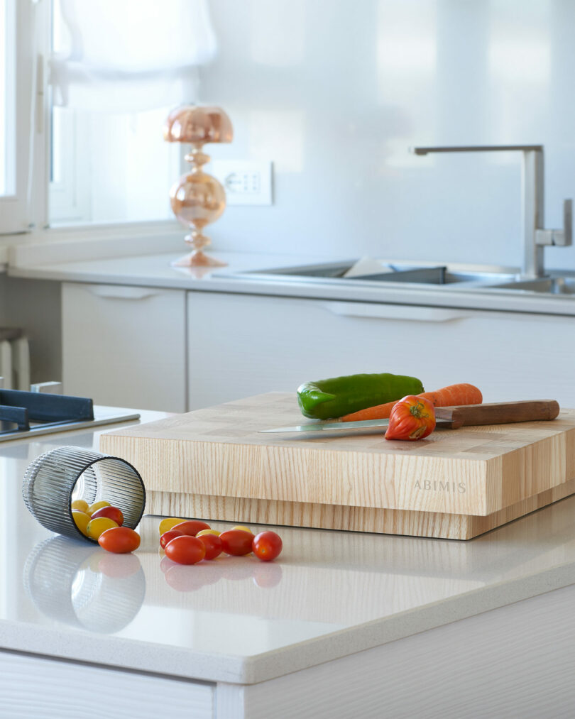 A kitchen countertop with a wooden cutting board holding a knife, green pepper, and carrot; nearby are a wire basket and scattered cherry tomatoes