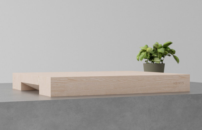 A light wood monitor stand on a gray surface with a small potted plant in the background