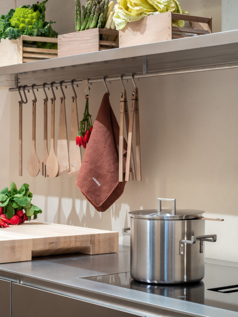 A stainless steel pot with a lid sits on a stovetop; wooden utensils and a brown towel hang above, with crates of vegetables on a shelf and a cutting board with greens nearby