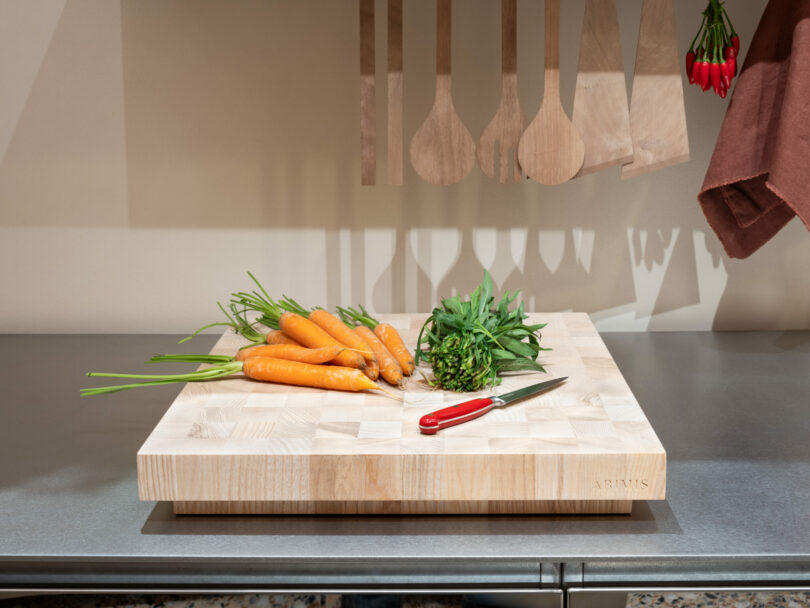 A wooden cutting board with whole carrots, carrot greens, and a red-handled knife on a stainless steel countertop; wooden utensils hang in the background.