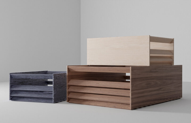 Three wooden crates of varying sizes and shades—dark grey, medium brown, and light beige—are stacked in a minimalist, light-colored room