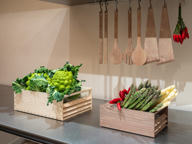 Two wooden crates on a kitchen counter hold Romanesco broccoli, leafy greens, green asparagus, white asparagus, and red chili peppers; wooden utensils hang above