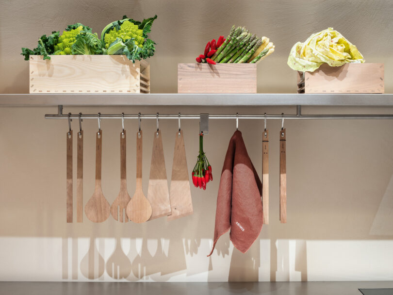Three wooden crates with vegetables sit on a shelf above a rail holding wooden kitchen utensils, a bunch of red chili peppers, and a pink dish towel