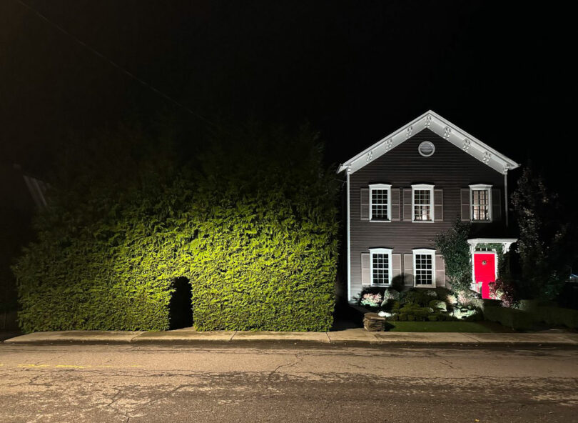 A two-story house with a red front door is next to a large hedge, which has a small archway cut into it; the scene is illuminated at night.