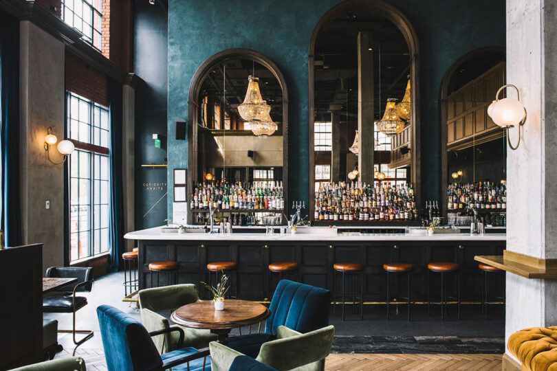 A modern bar with a long counter, brown leather stools, large mirrors, chandeliers, and stylish lounge seating in front of tall windows.