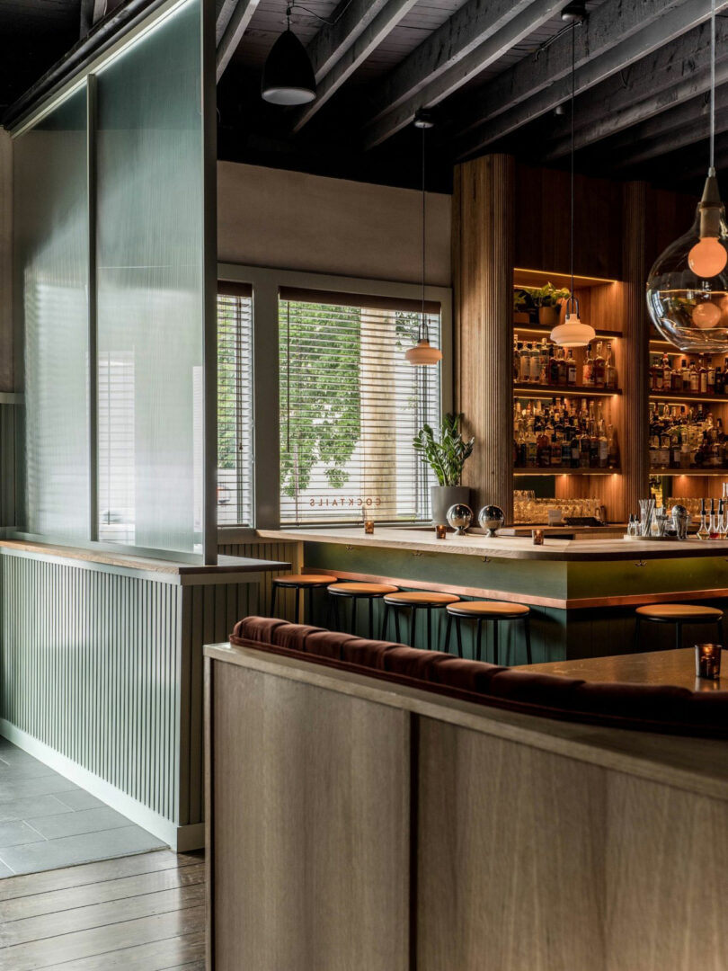 Modern bar interior featuring wooden furniture, a stocked backlit bar, pendant lights, and large windows with blinds letting in natural light.