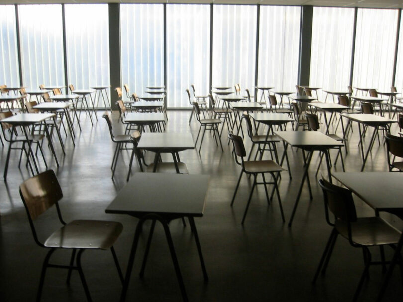 Rows of empty desks and chairs are arranged in a spacious, sunlit classroom with large windows and a polished floor.