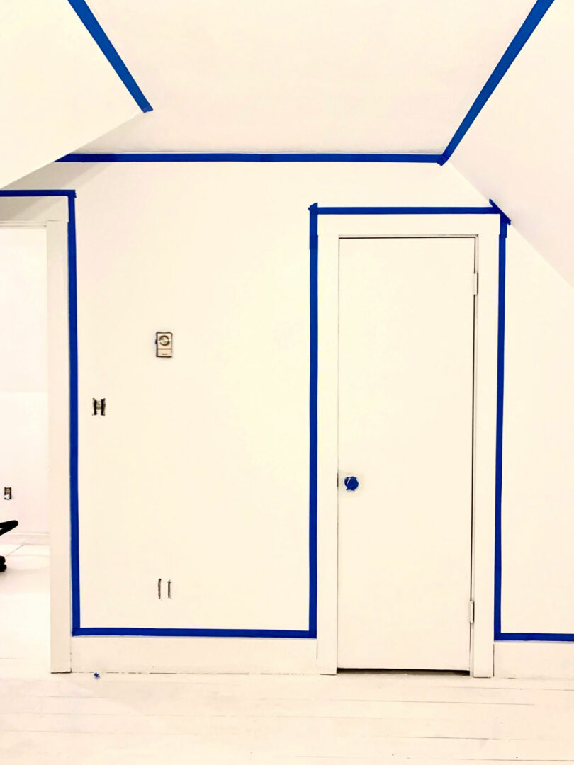 A white room with blue painter’s tape outlining the walls, ceiling edges, door, and trim in preparation for painting.