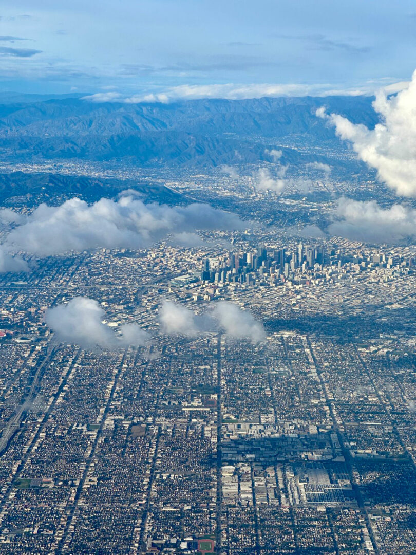Aerial view of a densely populated urban area with a cluster of skyscrapers near the center, mountains in the background, and scattered clouds above the city.
