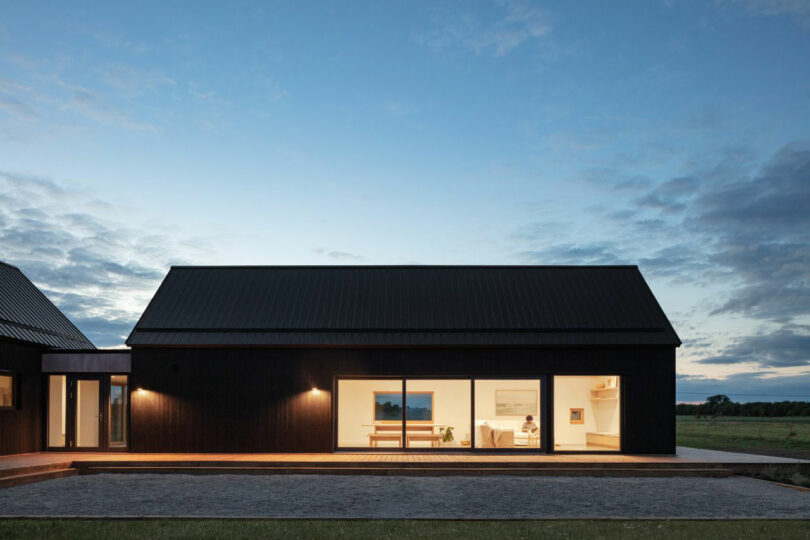 A modern, single-story house with dark exterior walls, large windows, and interior lights on, set against an evening sky with clouds and open landscape in the background.