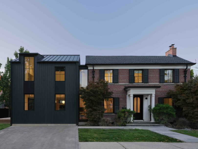 Two-story house with a modern black vertical siding extension on the left and a traditional red brick section on the right, featuring black shutters and illuminated windows.