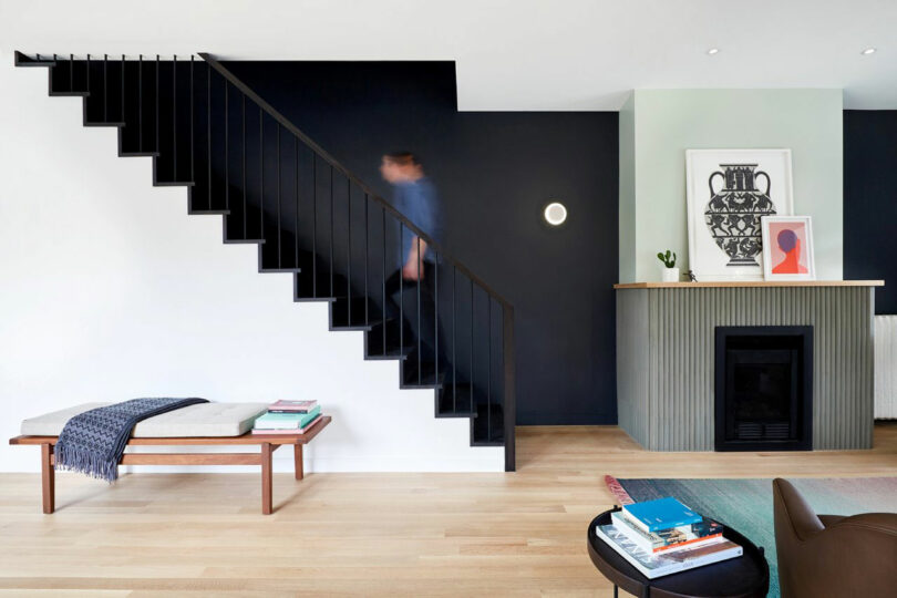 A person walks up a black staircase in a modern living space with a bench, fireplace, and minimal decor.