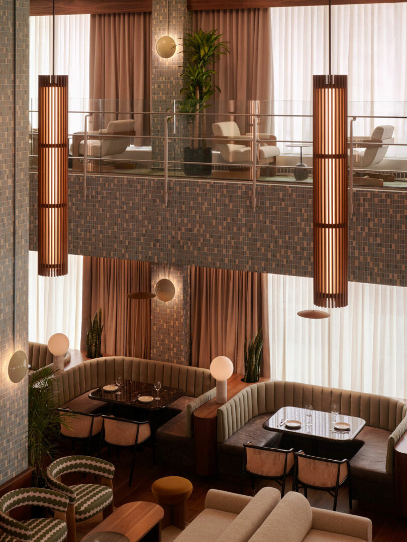 A modern, two-level restaurant interior features beige and brown tones, mosaic walls, round pendant lights, and upholstered booth seating with tables set for dining.