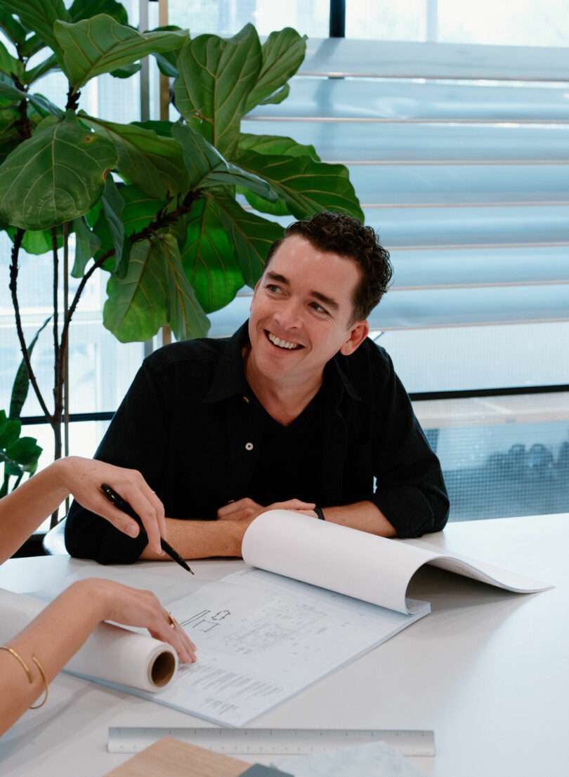 A man sits at a table smiling, looking at someone off-camera, with architectural plans and a rolled-up document in front of him. A large plant is in the background.