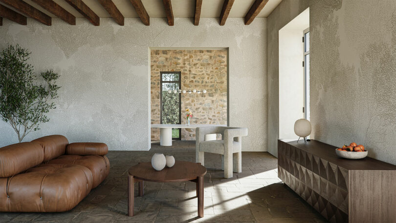 Modern living room with a brown leather sofa, round wooden coffee table, white armchair, sideboard with a lamp and fruit bowl, and textured stone walls with wooden ceiling beams