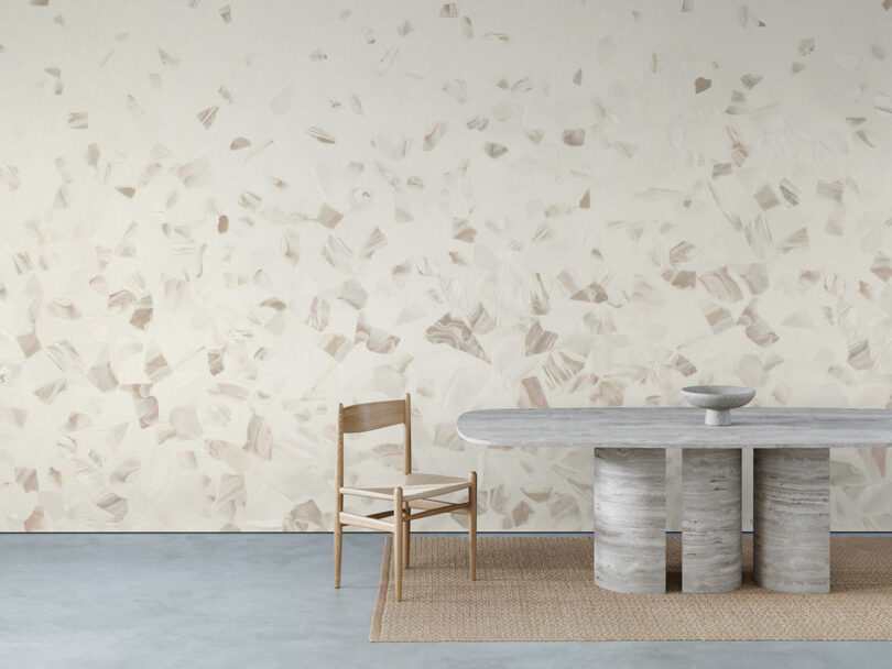 Minimalist dining area with a stone table, a single wooden chair, a woven rug, and a abalone-patterned wall in neutral tones.