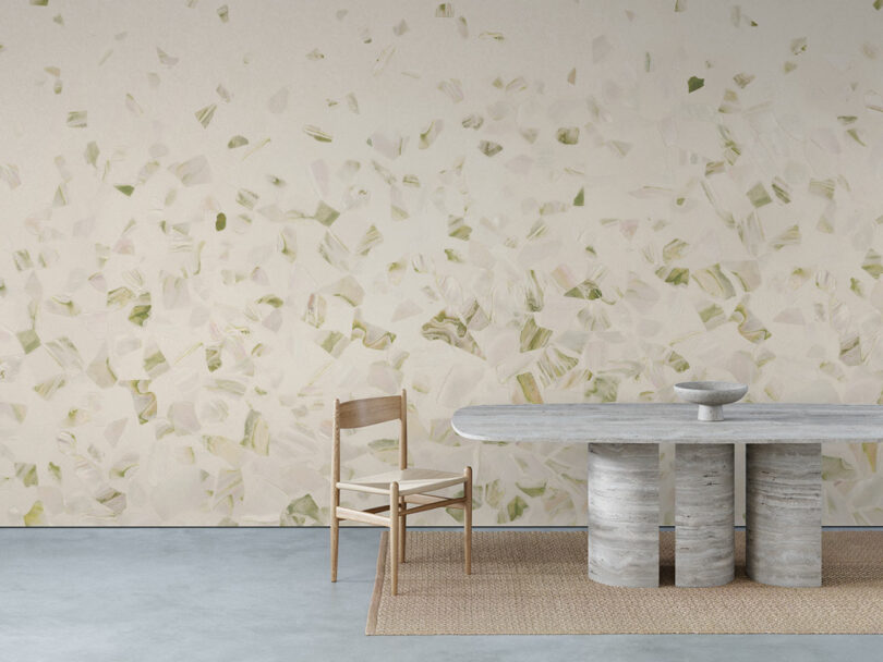 A minimalist room with a abalone-patterned wall, a stone table with three cylindrical legs, a wooden chair, a bowl, and a woven rug on a concrete floor.
