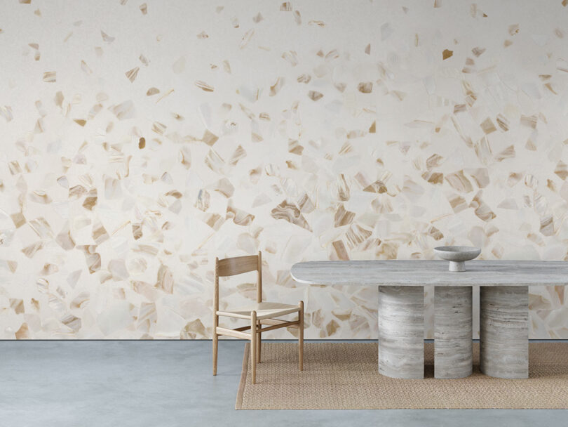 A minimalist room with a abalone-patterned wall, a wooden chair, a stone table with three cylindrical legs, and a woven rug on a concrete floor.