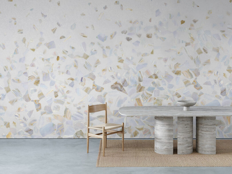 A minimalist dining area with a wooden chair, a stone table on a woven rug, and a wall featuring a pastel abalone pattern
