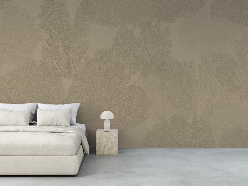 Minimalist bedroom with a beige upholstered bed, white bedding, a small stone side table, white lamp, and a taupe wall featuring a subtle botanical pattern