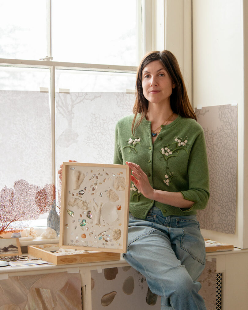 A woman in a green sweater sits by a window, holding and displaying a framed collection of seashells and natural objects