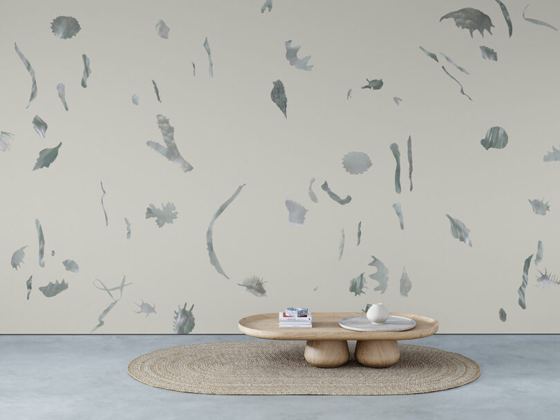 A minimalist room with a round woven rug, a low wooden coffee table with books and a vase, and a wall decorated with abstract grayish patterns.