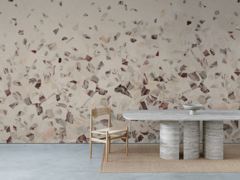 A minimalist room with a abalone-patterned wall, a wooden chair, a stone table with cylindrical legs, and a bowl, all set on a woven rug over a concrete floor