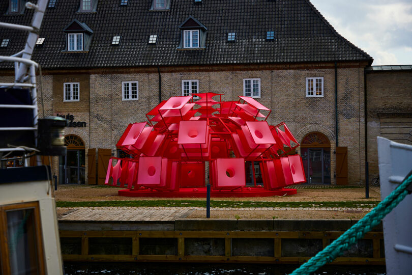 A large red geometric sculpture made of square panels is displayed on grass in front of a brick building with a dark tile roof near a waterfront.