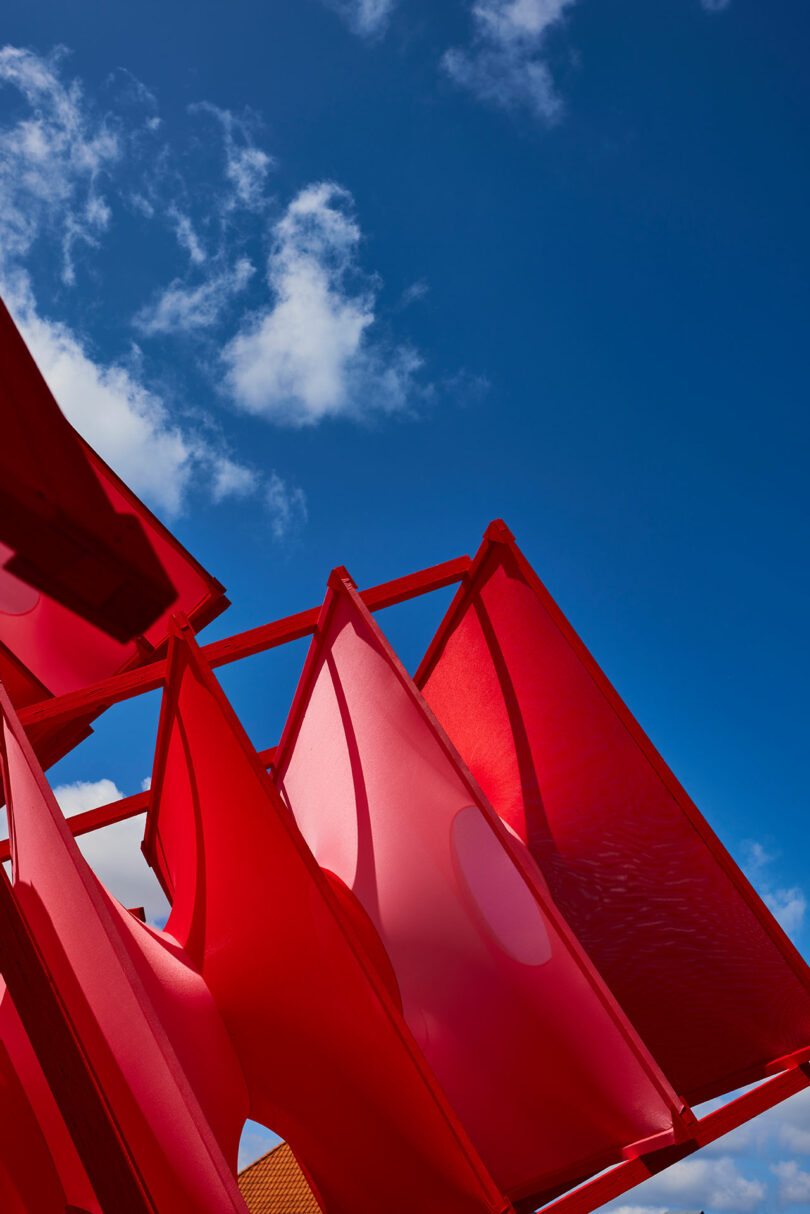 Red geometric fabric panels are arranged diagonally against a bright blue sky with scattered clouds.