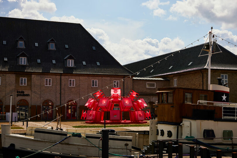 A cluster of bright red modular structures is set up on a grassy area between two brick buildings near a harbor with docked boats.