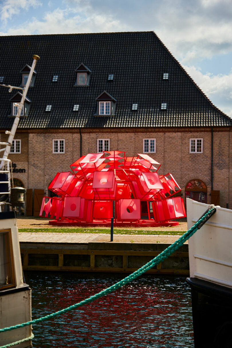 A red, dome-shaped art installation made of panels stands on grass in front of a brick building with dormer windows by a canal.