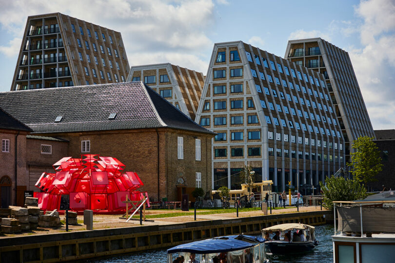 A canal scene with modern, angular glass buildings, a traditional brick structure, a cluster of red geometric sculptures, and boats on the water under a partly cloudy sky.