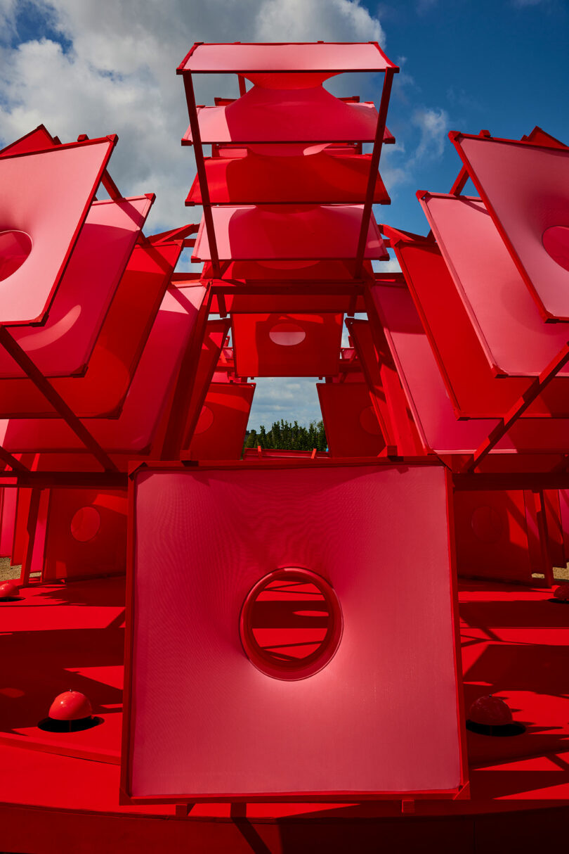 Large geometric sculpture made of stacked red square panels with central circular cutouts, set outdoors under a partly cloudy sky.