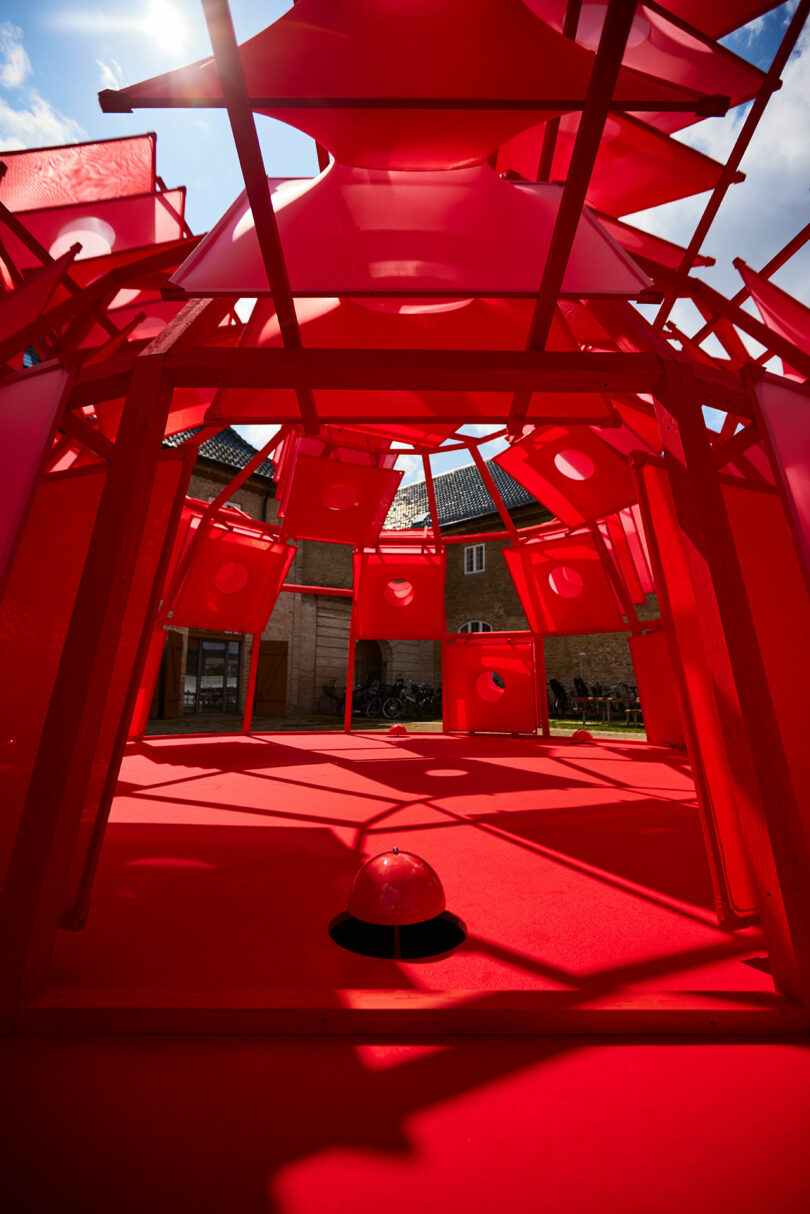 Interior view of a geometric red art installation with circular cut-outs, beneath a sunny sky and in front of a brick building.