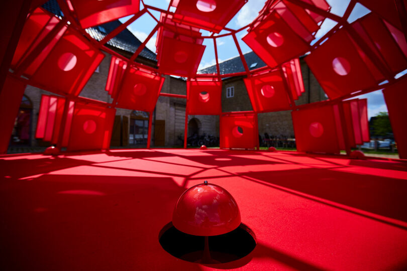 Red geometric art installation with square panels and circular cutouts, viewed from inside. A red dome structure sits on the floor, with buildings visible through openings outside.