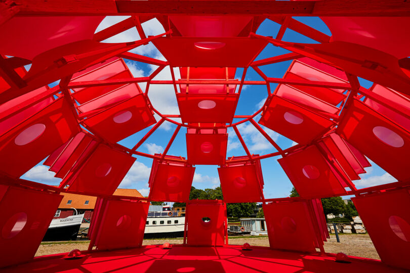 View from inside a bright red geometric structure with circular openings, looking out towards buildings, trees, and a partly cloudy sky.