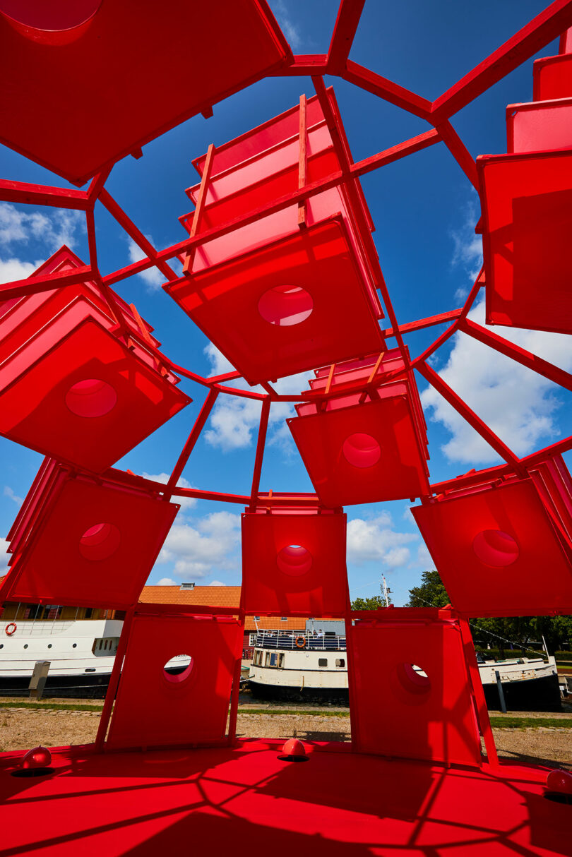 Red geometric structure with circular cutouts seen from inside, set against a blue sky with clouds; white boats and a building are visible in the background.