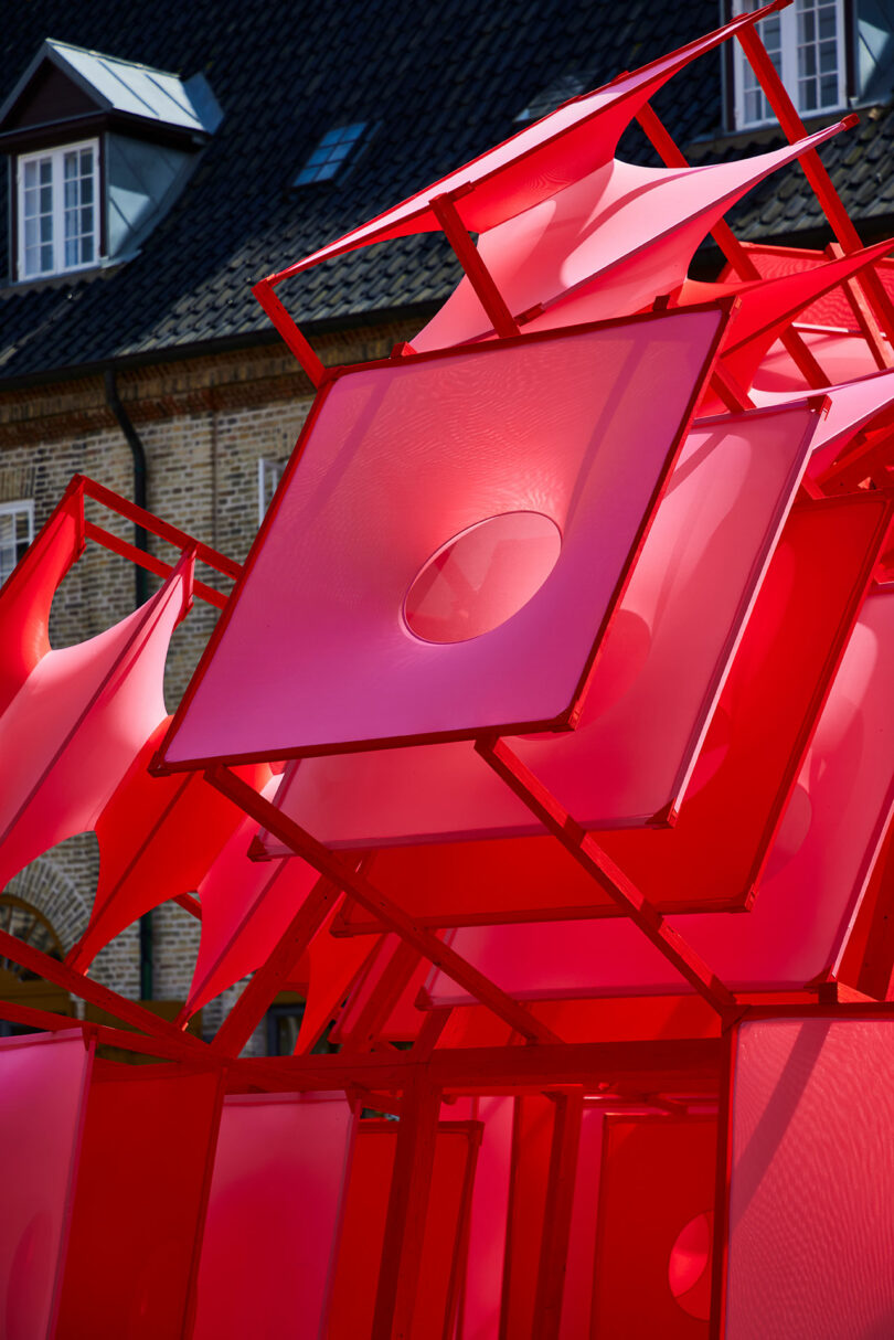 A close-up of a modern outdoor sculpture made of translucent red geometric panels, set against a brick building with windows and a tiled roof.